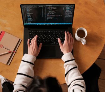 Top-down view of a person typing on a laptop displaying code, with a notebook, pen, and coffee cup on a wooden table